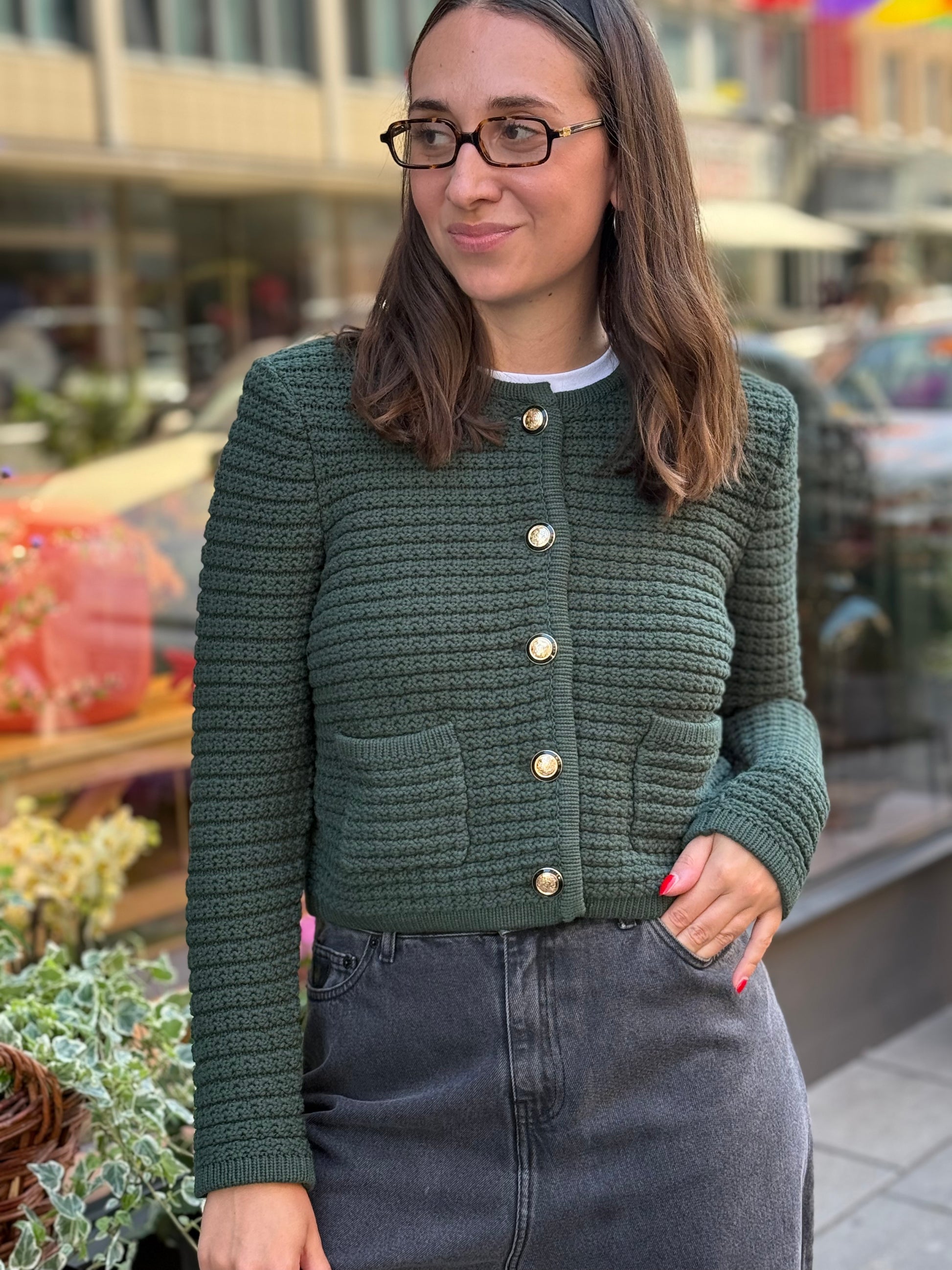 Frau steht vor Blumenladen, sie trägt einen grünen Cardigan mit goldenen Knöpfen und eine graue Jeans.