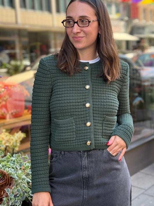 Frau steht vor Blumenladen, sie trägt einen grünen Cardigan mit goldenen Knöpfen und eine graue Jeans.