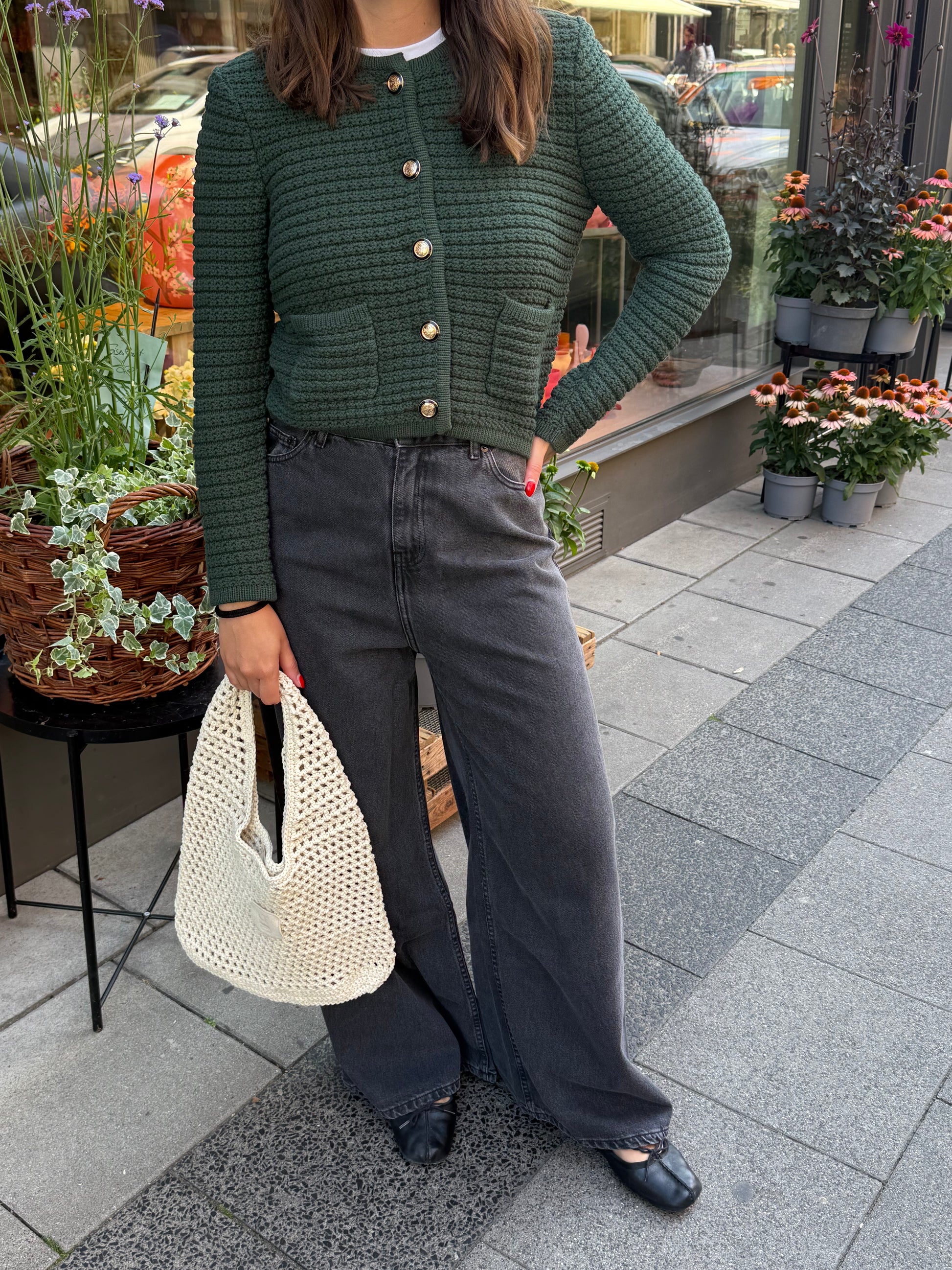 Frau steht vor Blumenladen, sie trägt einen grünen Cardigan mit goldenen Knöpfen und eine graue Jeans.