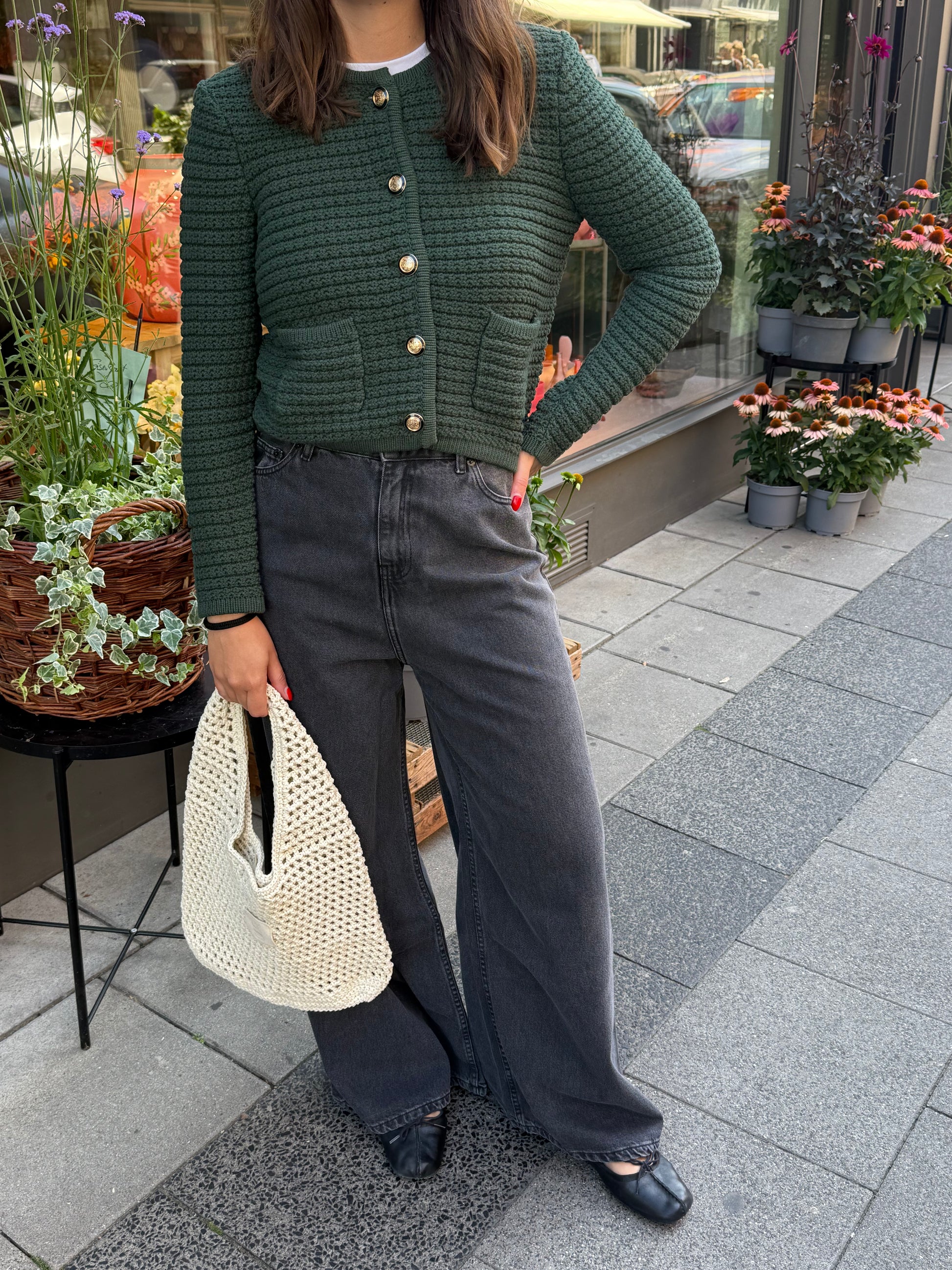 Frau steht vor Blumenladen, sie trägt eine graue weite Denim und einen waldgrünen ardigan mit goldenen Knöpfen. 
