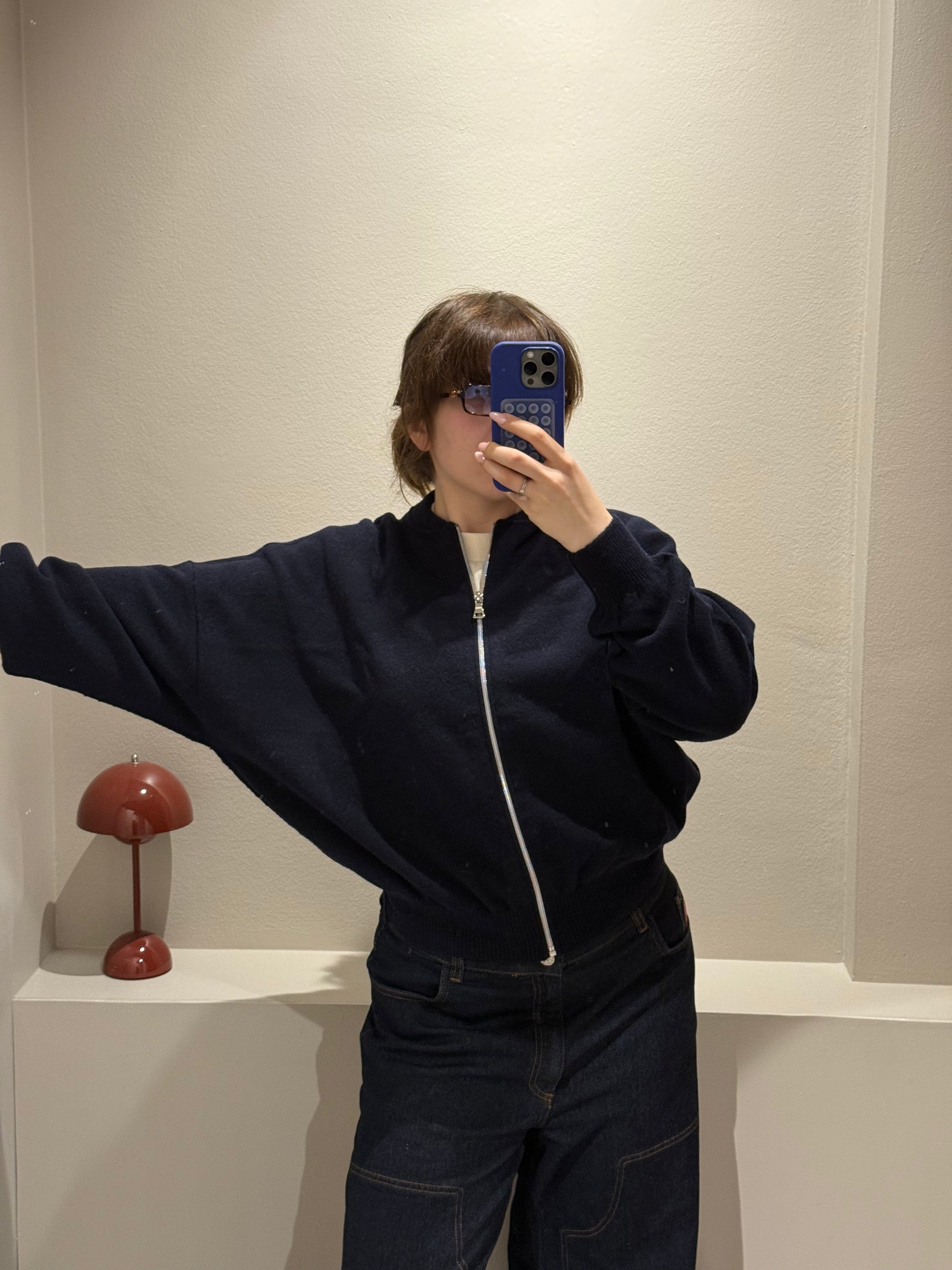 Frau macht Spiegelselfie, sie trägt iene dunkelblaue Strickbomberjacke und dunkle Jeans.