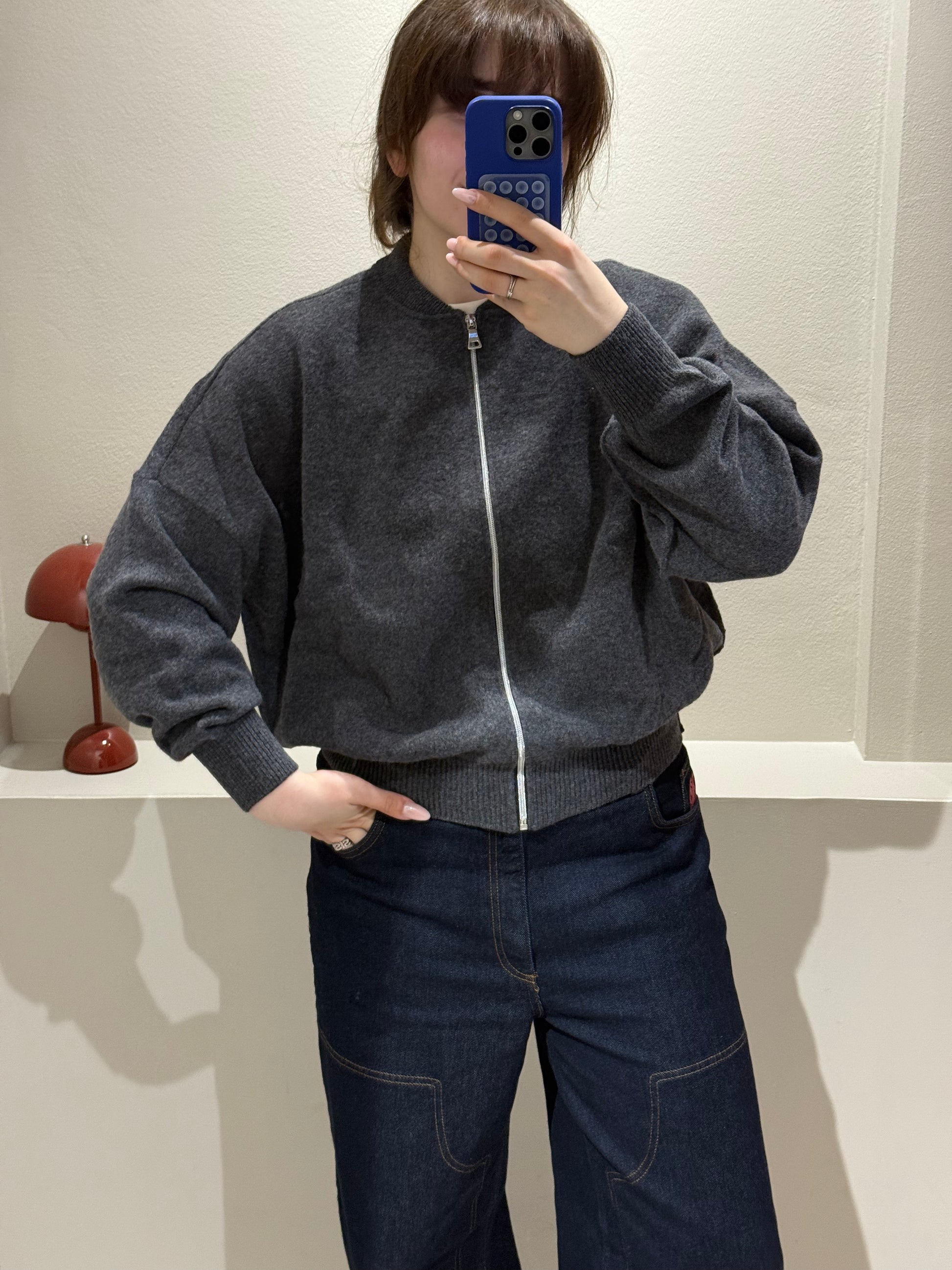 Frau macht Spiegelselfie, sie trägt iene dunkelgraue Strickbomberjacke und dunkle Jeans.