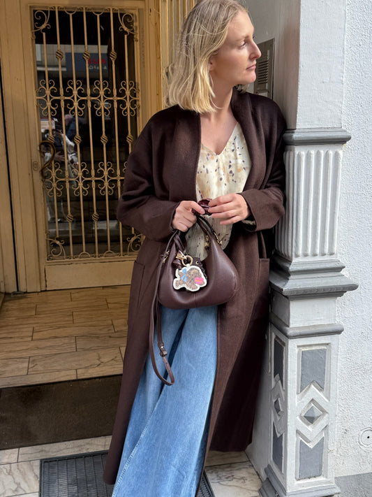 Frau steht in einem Türeingang sie trägt einen braunen Mantel, Jenas und bluse, dazu eine braune Tasche.