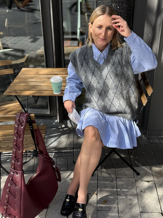Frau sitz an einem Outdoor Cafe Tisch sie trägt ien blaues Kleid und darüber eienn grauen Pullunder mit Karo Muster. Es ist eine klassiche Cafe Situation.