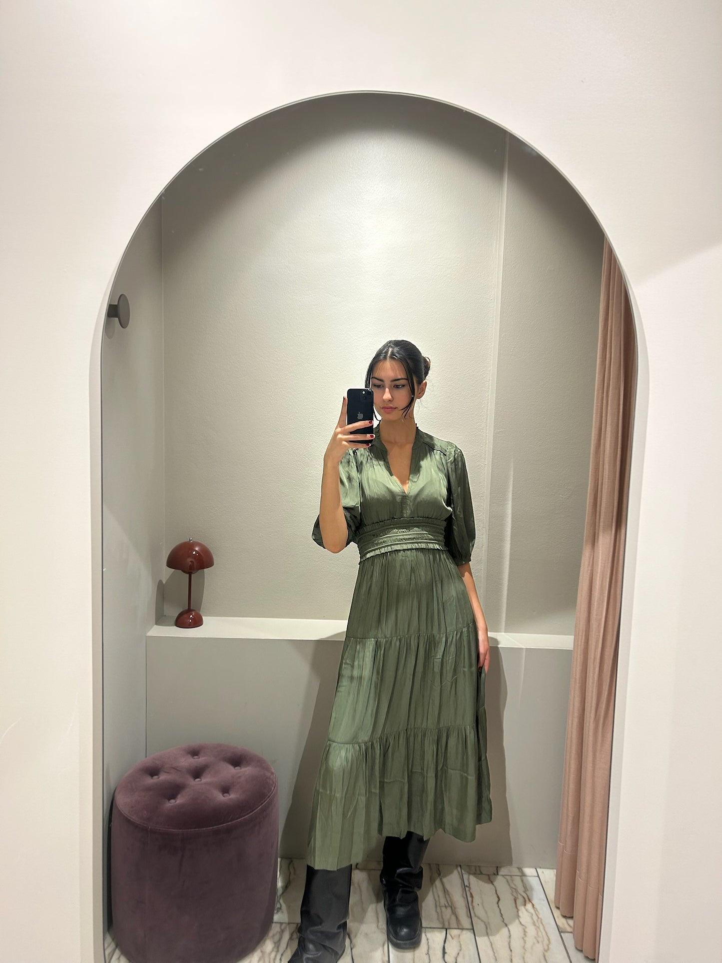Frau trägt ein wadenlanges Kleid in Olivgrün mit V-Ausschnitt, halblangen Ärmeln und Stufenrock. Das Kleid hat eine betonte Taille und einen weich fließenden Stoff. Sie kombiniert es mit schwarzen Stiefeln und macht ein Spiegelselfie in einer Umkleidekabine mit hellem Bogenrahmen.