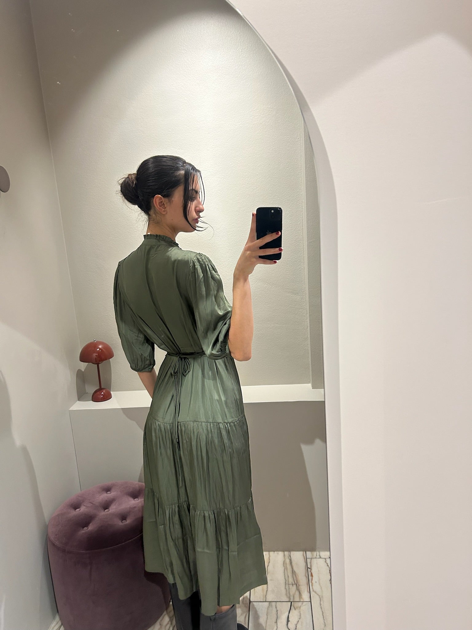 Frau trägt ein wadenlanges Kleid in Olivgrün mit V-Ausschnitt, halblangen Ärmeln und Stufenrock. Das Kleid hat eine betonte Taille und einen weich fließenden Stoff. Sie kombiniert es mit schwarzen Stiefeln und macht ein Spiegelselfie in einer Umkleidekabine mit hellem Bogenrahmen.