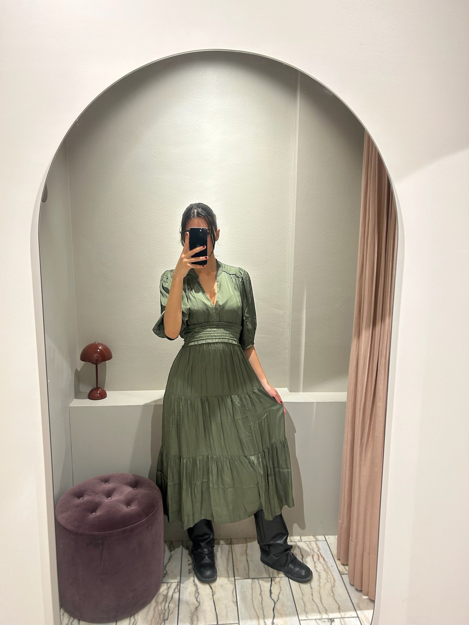 Frau trägt ein wadenlanges Kleid in Olivgrün mit V-Ausschnitt, halblangen Ärmeln und Stufenrock. Das Kleid hat eine betonte Taille und einen weich fließenden Stoff. Sie kombiniert es mit schwarzen Stiefeln und macht ein Spiegelselfie in einer Umkleidekabine mit hellem Bogenrahmen.