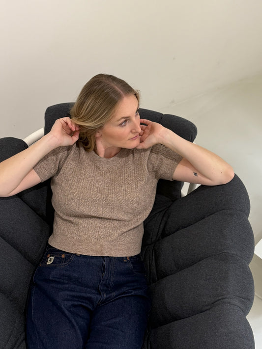 Frau sitzt auf Sessel, sie trägt braunes Strick Tee und dunkle Jeans.