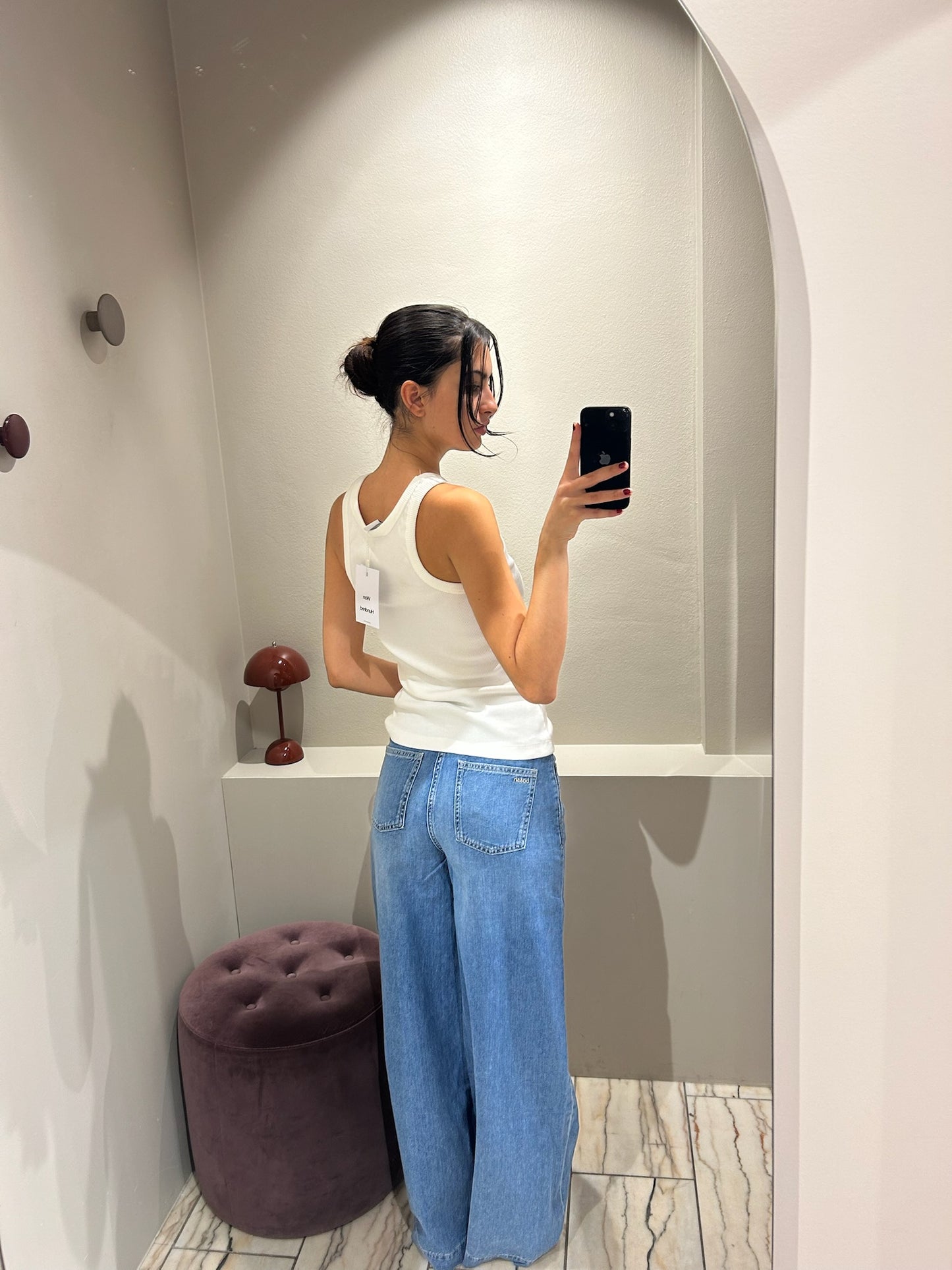 Frau steht in einer Umkleidekabine und trägt eine hellblaue Jeans mit weitem Bein und mittelhohem Bund. Dazu kombiniert sie ein weißes, ärmelloses Top mit Rundhalsausschnitt. Sie macht ein Spiegelselfie vor einem hellen, gebogenen Wandspiegel. Der Hintergrund ist minimalistisch gehalten mit beiger Wand, Vorhang und einem runden, violetten Hocker.