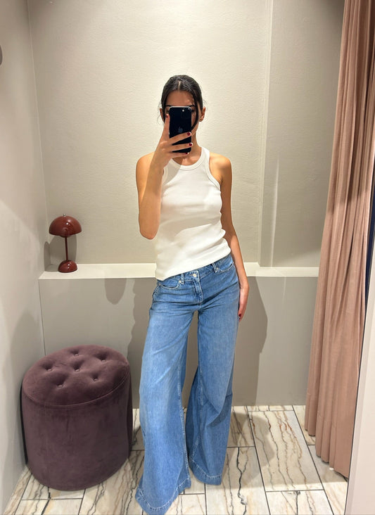 Frau steht in einer Umkleidekabine und trägt eine hellblaue Jeans mit weitem Bein und mittelhohem Bund. Dazu kombiniert sie ein weißes, ärmelloses Top mit Rundhalsausschnitt. Sie macht ein Spiegelselfie vor einem hellen, gebogenen Wandspiegel. Der Hintergrund ist minimalistisch gehalten mit beiger Wand, Vorhang und einem runden, violetten Hocker.