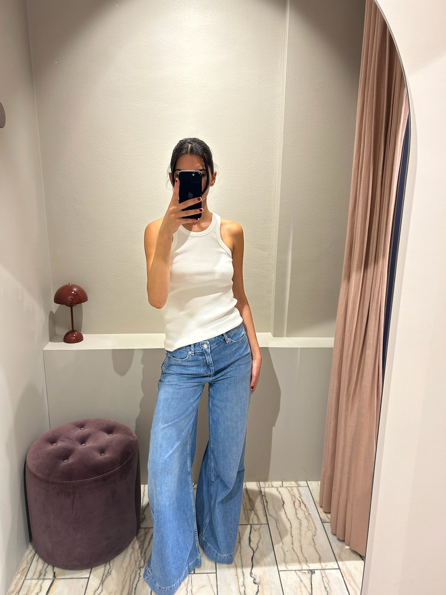Frau steht in einer Umkleidekabine und trägt eine hellblaue Jeans mit weitem Bein und mittelhohem Bund. Dazu kombiniert sie ein weißes, ärmelloses Top mit Rundhalsausschnitt. Sie macht ein Spiegelselfie vor einem hellen, gebogenen Wandspiegel. Der Hintergrund ist minimalistisch gehalten mit beiger Wand, Vorhang und einem runden, violetten Hocker.