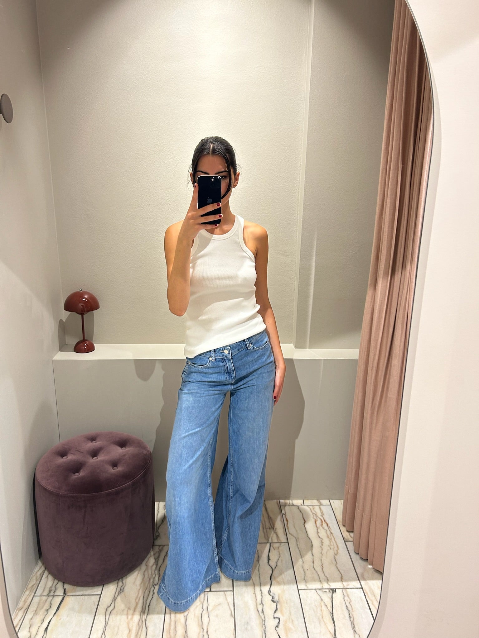 Frau steht in einer Umkleidekabine und trägt eine hellblaue Jeans mit weitem Bein und mittelhohem Bund. Dazu kombiniert sie ein weißes, ärmelloses Top mit Rundhalsausschnitt. Sie macht ein Spiegelselfie vor einem hellen, gebogenen Wandspiegel. Der Hintergrund ist minimalistisch gehalten mit beiger Wand, Vorhang und einem runden, violetten Hocker.