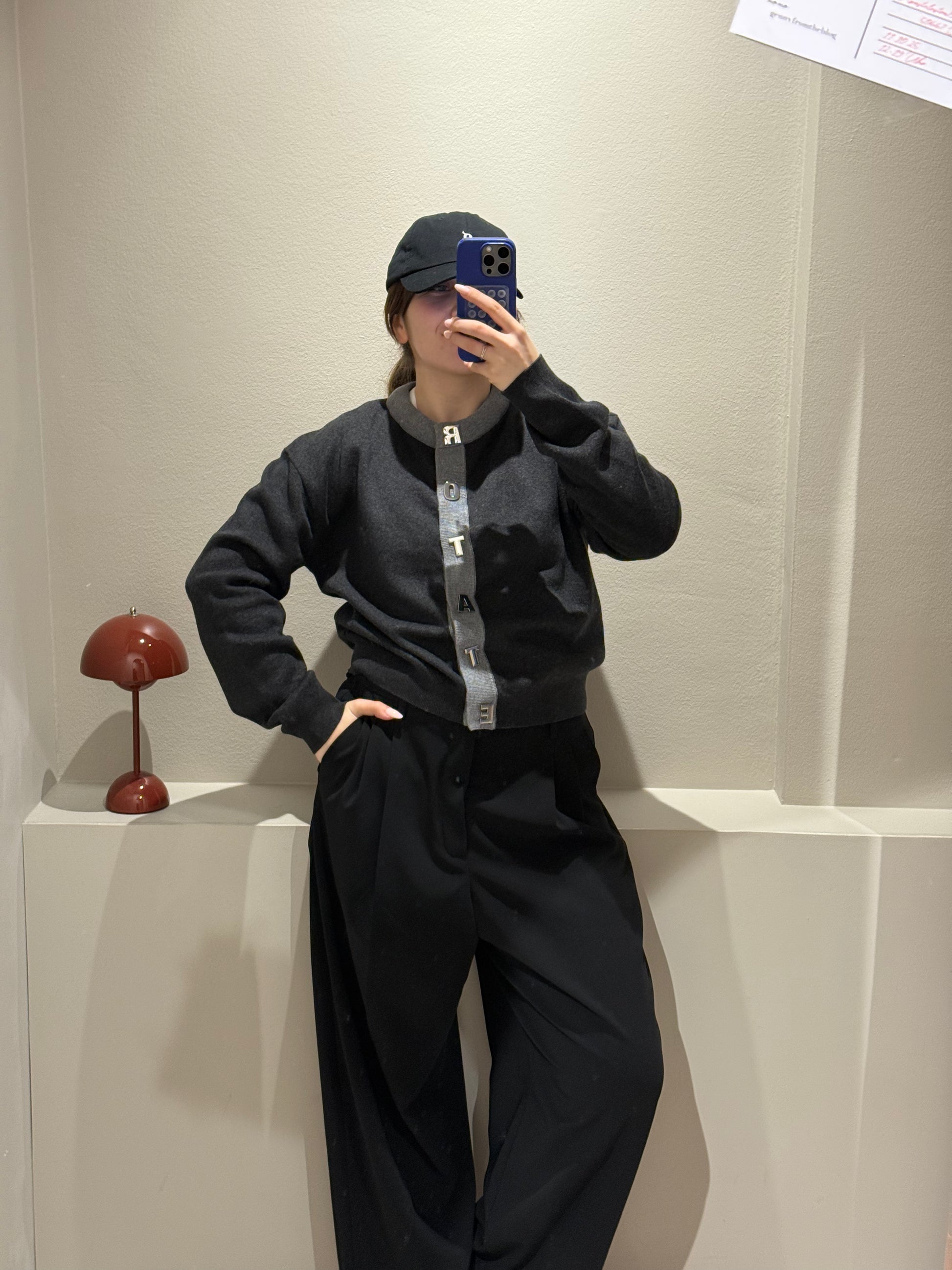 Frau macht Spiegelselfie, sie trägt eine schwarze Hose und einen grauen Cardigan mit Kontrastsäumen und Rotate geschrieben an der Knopfleiste.