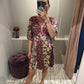 Frau steht in einer Umkleidekabine und macht ein Spiegelselfie. Sie trägt ein geblümtes, bordeauxfarbenes Kleid mit V-Ausschnitt, kurzem Schnitt und halblangen Ärmeln. Das Kleid hat ein Patchwork-Muster mit floralen Prints in Rosa, Creme und Grün sowie pinkfarbene Zierpaspeln. Der leicht ausgestellte Rock fällt locker über einen sichtbaren Babybauch. Die Frau trägt schwarze Zehensandalen und hat ihre blonden Haare streng zurückgebunden