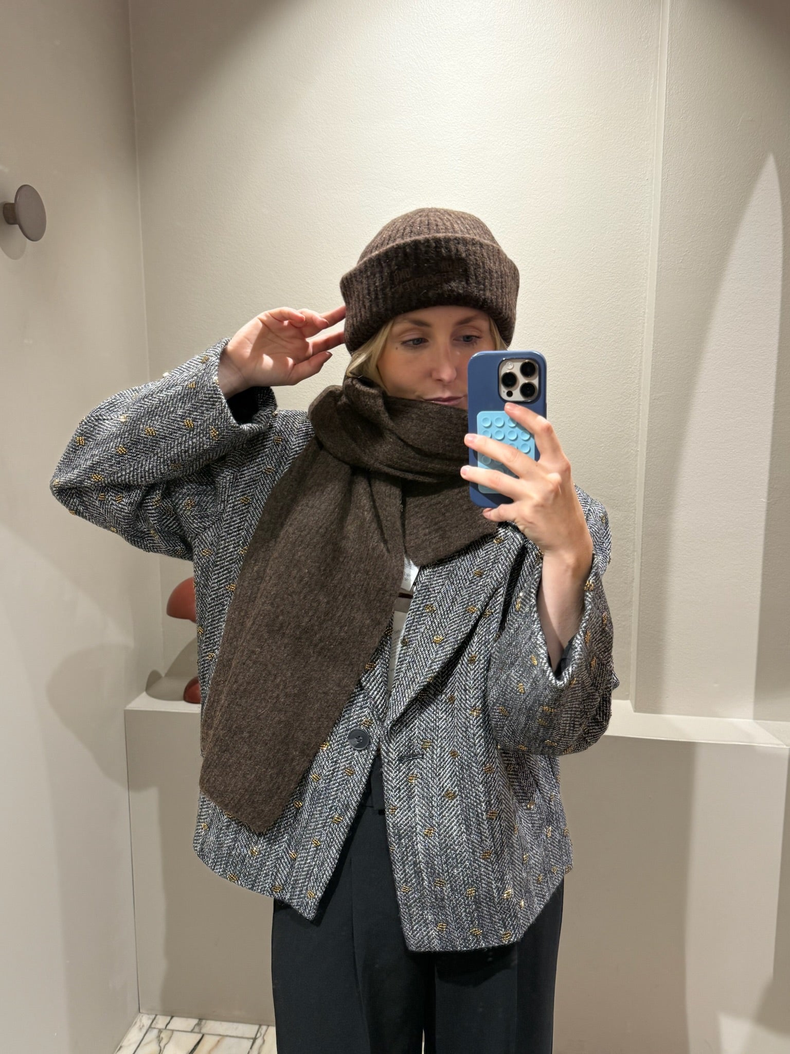 Frau macht Spiegelselfie, sie trägt eine schwarze Hose dazu einen grauen Blazer mit Pailletten und braune Mütze und Schal.