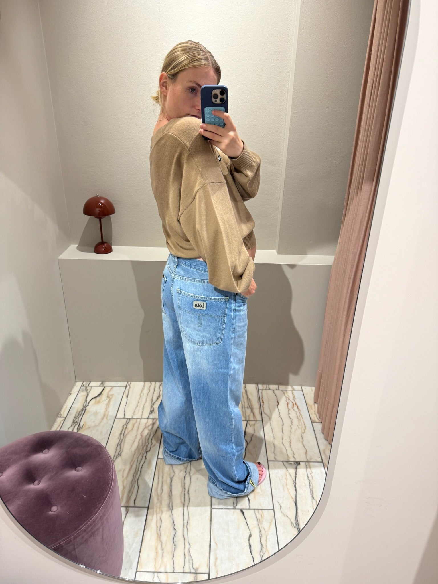 Frau macht Spiegelselfie, sie trägt eine Weite hellblaue Jeans und einen Camelfarbenden V-Neck Strick. Das Fote zeigt die Jeans von hinten, man sieht die taschen am Po.
