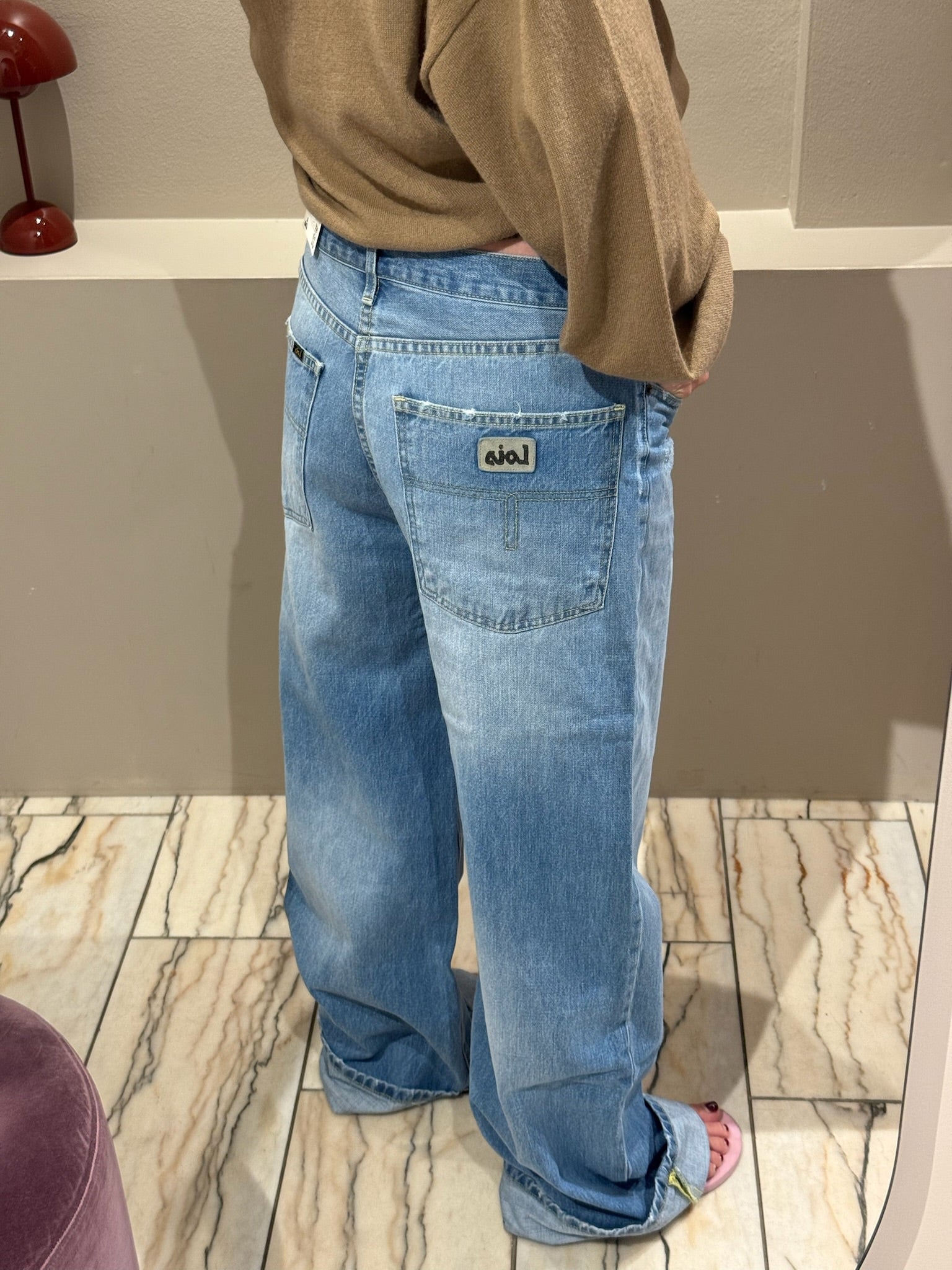 Frau macht Spiegelselfie, sie trägt eine Weite hellblaue Jeans und einen Camelfarbenden V-Neck Strick. Das Fote zeigt die Jeans von hinten, man sieht die taschen am Po.