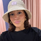 Frau macht ein Selfie vor rosa Vorhängen. Sie trägt einen naturfarbenen Strohhut im Bucket-Hat-Stil mit einem lila Aufnäher, auf dem ein gesticktes ‚g‘ zu sehen ist. Dazu trägt sie ein schwarzes Oberteil und goldene Creolen. Sie blickt direkt in die Kamera und lächelt leicht.
