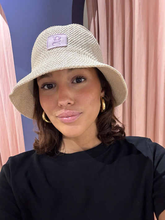 Frau macht ein Selfie vor rosa Vorhängen. Sie trägt einen naturfarbenen Strohhut im Bucket-Hat-Stil mit einem lila Aufnäher, auf dem ein gesticktes ‚g‘ zu sehen ist. Dazu trägt sie ein schwarzes Oberteil und goldene Creolen. Sie blickt direkt in die Kamera und lächelt leicht.
