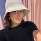 Frau macht ein Selfie vor rosa Vorhängen. Sie trägt einen naturfarbenen Strohhut im Bucket-Hat-Stil mit einem lila Aufnäher, auf dem ein gesticktes ‚g‘ zu sehen ist. Dazu trägt sie ein schwarzes Oberteil und goldene Creolen. Sie blickt direkt in die Kamera und lächelt leicht.