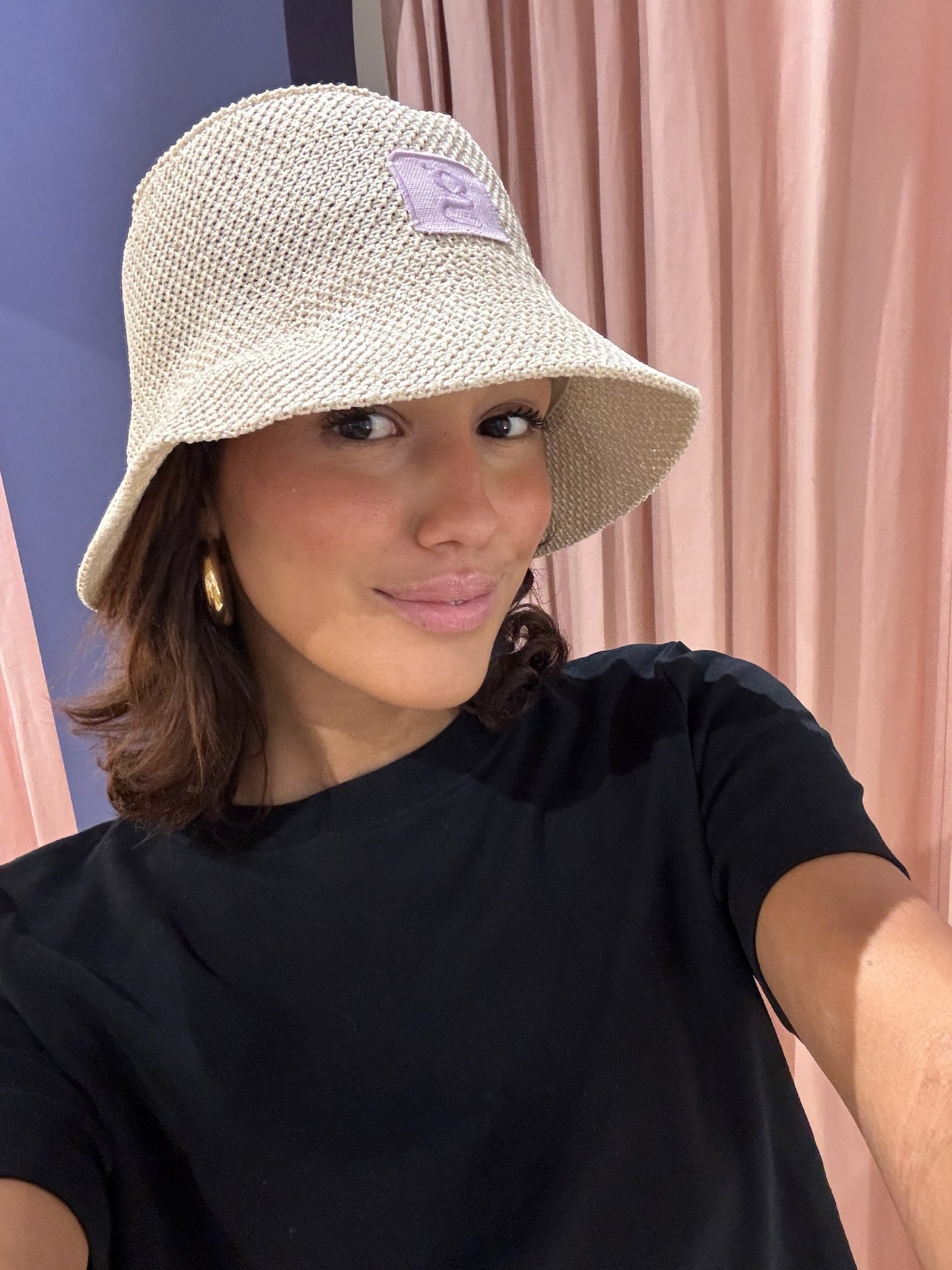 Frau macht ein Selfie vor rosa Vorhängen. Sie trägt einen naturfarbenen Strohhut im Bucket-Hat-Stil mit einem lila Aufnäher, auf dem ein gesticktes ‚g‘ zu sehen ist. Dazu trägt sie ein schwarzes Oberteil und goldene Creolen. Sie blickt direkt in die Kamera und lächelt leicht.