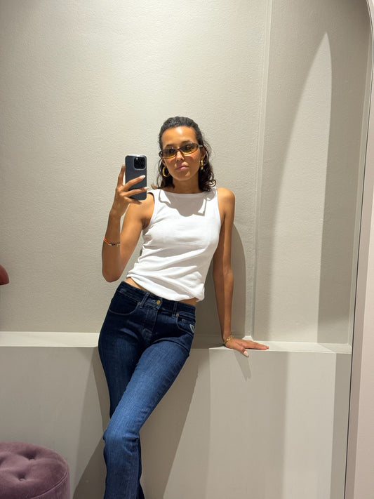 Frau macht Spiegelselfie, sie trägt ein weißes Takntop und eine dunkleblaue Jeans mit Bootcut. Sie lehnt sich an einer Wand ab.