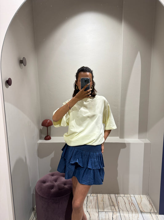 ine Frau steht in einer Umkleidekabine und macht ein Selfie vor einem Spiegel. Sie trägt ein oversized T-Shirt in zartem Buttergelb mit kurzen Ärmeln. Auf der linken Brust ist ein gesticktes ‚G‘ zu sehen, am linken Ärmel eine kleine Zitronen-Stickerei. Dazu kombiniert sie einen kurzen, dunkelblauen Stufenrock mit gesmoktem Bund und Volants. Ihr Gesicht ist größtenteils vom Smartphone verdeckt
