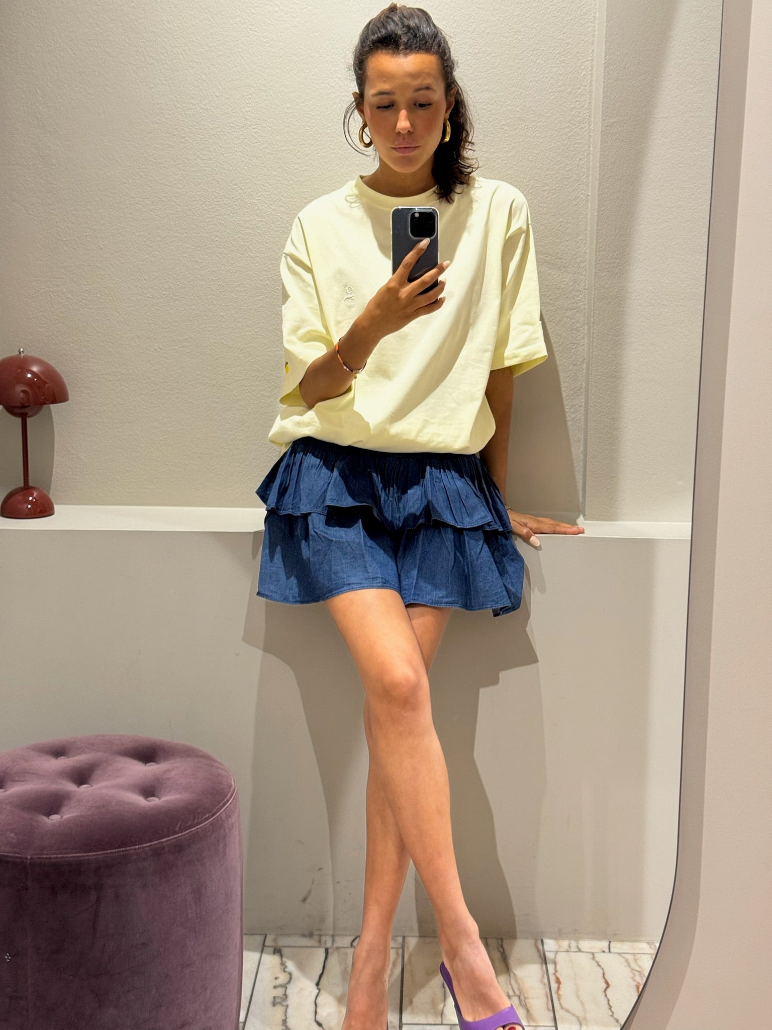 ine Frau steht in einer Umkleidekabine und macht ein Selfie vor einem Spiegel. Sie trägt ein oversized T-Shirt in zartem Buttergelb mit kurzen Ärmeln. Auf der linken Brust ist ein gesticktes ‚G‘ zu sehen, am linken Ärmel eine kleine Zitronen-Stickerei. Dazu kombiniert sie einen kurzen, dunkelblauen Stufenrock mit gesmoktem Bund und Volants. Ihr Gesicht ist größtenteils vom Smartphone verdeckt