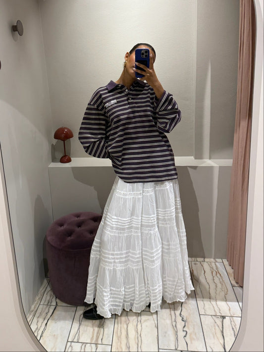 Eine Frau macht ein Spiegel Selfie, sie trägt einen weißen Rock mit horizontalen Lochstick-Streifen und Rüschensaum. Der rock ist sehr romantisch und fällt locker. Dazu styled sie ein oversized Polo und Cowboyboots.