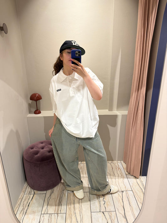 Eine Person steht in einer Umkleidekabine und macht ein Spiegel-Selfie. Sie trägt ein weißes, kurzärmeliges Polohemd mit klassischem Kragen und Knopfleiste. Auf der linken Brust befindet sich ein schwarzer Patch mit weißem Schriftzug ‚ROTATE‘.