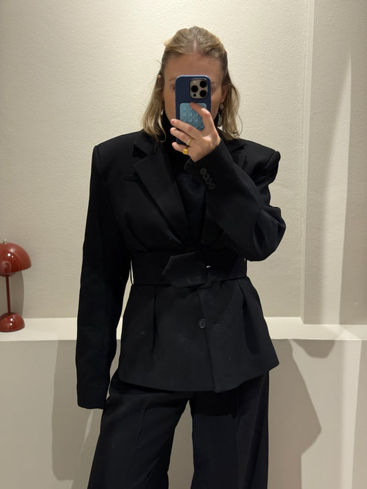Fraue macht Spigelselfie, sie trägt einen schwarzen taillierten Blazer mit schulterpolstern dazu schwarze Hose und Turtleneck.