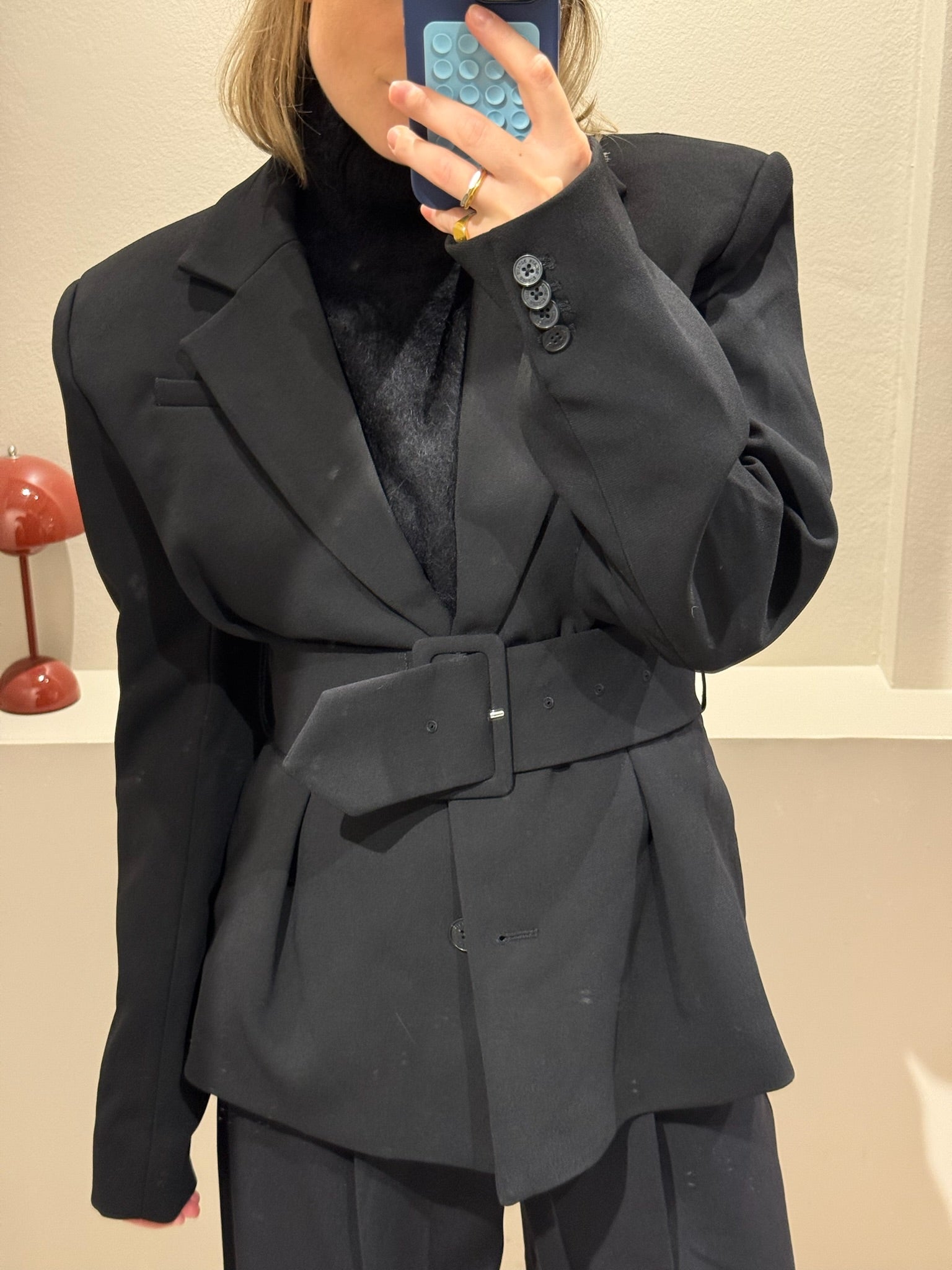 Fraue macht Spigelselfie, sie trägt einen schwarzen taillierten Blazer mit schulterpolstern dazu schwarze Hose und Turtleneck.
