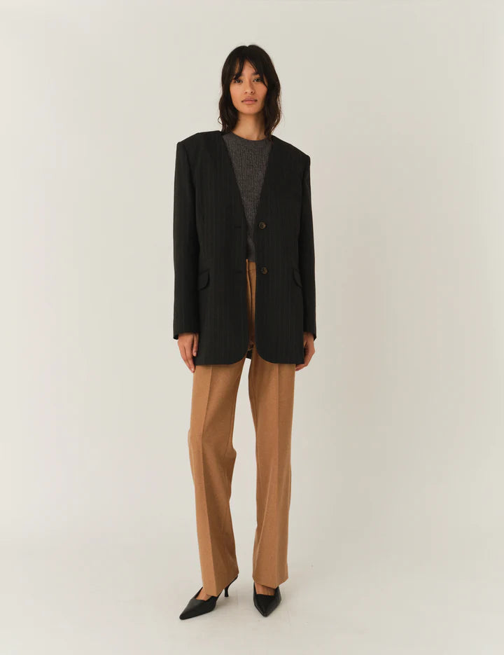 Frau vor weißem Hintergrund, sie trägt eine beige Hose und graues top Darüber einen Blazer ohne Reviere mit Nadelstreifen in dunkelgrau.