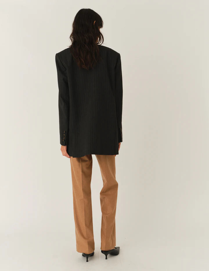 Frau vor weißem Hintergrund, sie trägt eine beige Hose und graues top Darüber einen Blazer ohne Reviere mit Nadelstreifen in dunkelgrau. Man sieht den Blazer von hinten.