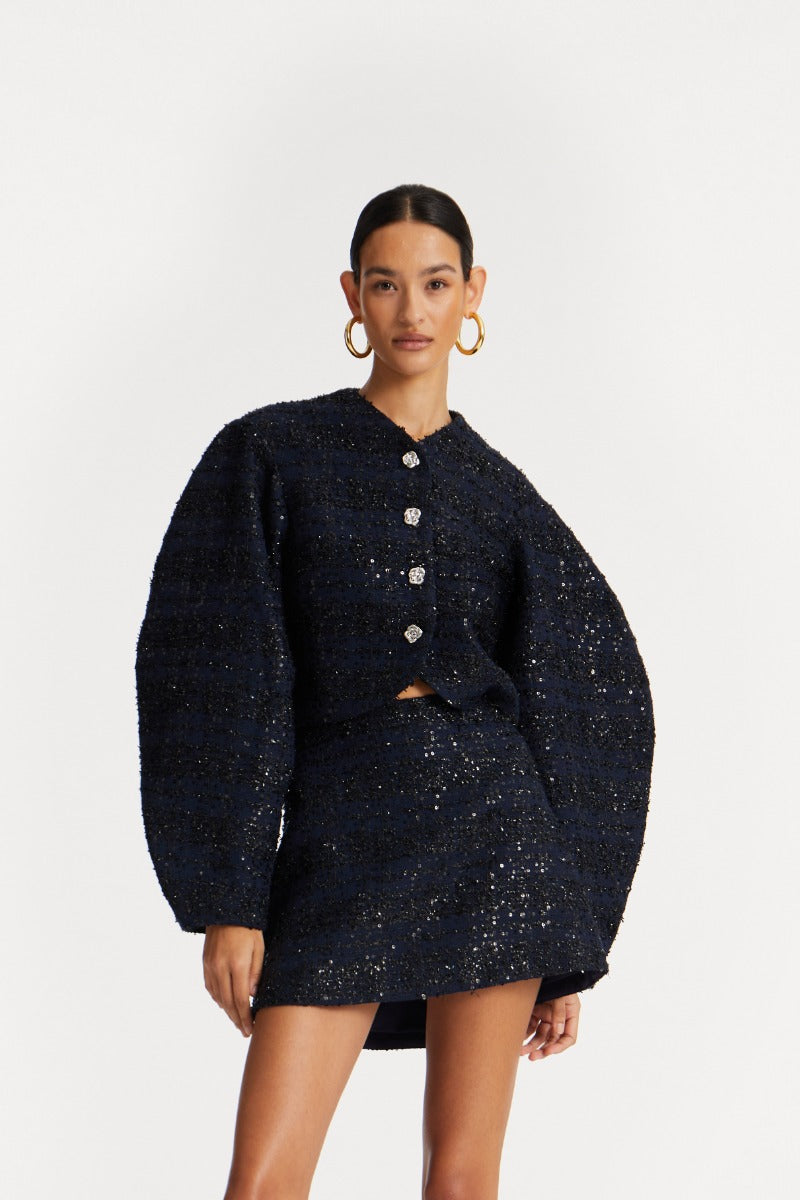 Frau vor weißem Backdrop. sie trägt eine dunkelblaue Boucle Jacke mit Pailliettebbesatz und passendem Minirock. Die Jacke ist leicht verkürzt und hat Ballonärmel.