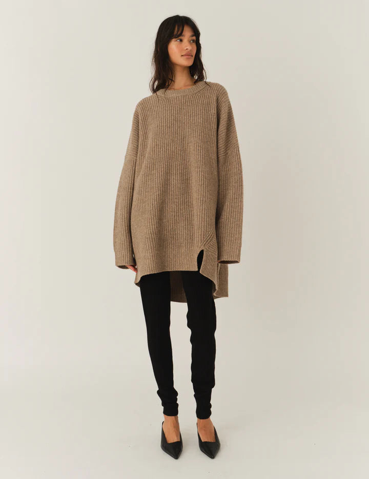Frau vor weißem Hintergrund, sie trägt einen beigen Oversized Strick über enger Hose und spitzen Schuhen.
